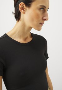 Juodas raukinis trumpas rankovėmis marškinėlis su apvaliu iškirpimu, turintis prigludusį dizainą ir lygų paviršių. Minimalistinis stilius be papuošimų.