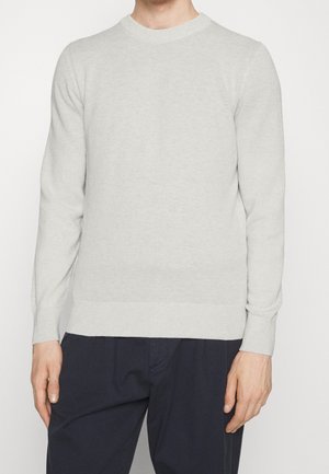 Man die een lichtgrijze gebreide trui met lange mouwen en een donkere broek draagt, staand tegen een effen witte achtergrond.