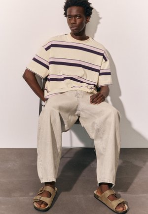 Ung mand, der sidder på en stol iført en stor beige t-shirt med striber, løstsiddende lyse jeans og beige sandaletter med spænder mod en ensfarvet baggrund.