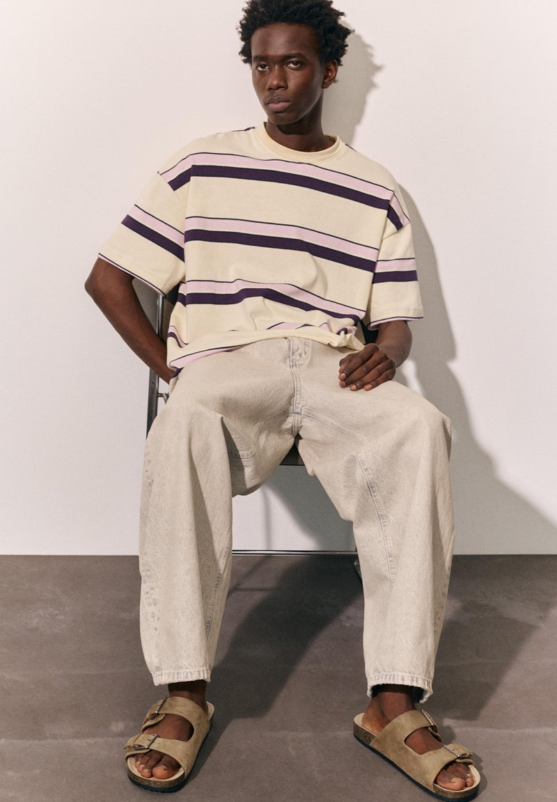 Jeune homme assis sur une chaise portant un t-shirt beige à rayures oversize, un jean clair ample et des sandales beiges à boucle sur fond uni.