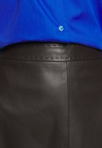 Jupe en cuir marron avec une texture lisse et des coutures discrètes. Associée à une chemise bleue ornée d'un détail de bouton décoratif.