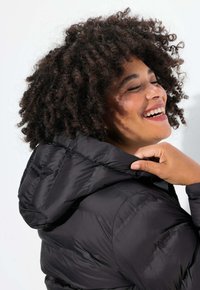 Abrigo de plumas negro con capucha, con textura acolchada, acabado elegante y cordón ajustable. El modelo ajusta el cuello.