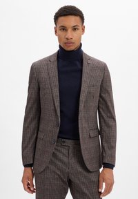 Braun kariertes Anzug mit einem strukturierten Blazer und passender Hose. Strukturierter Stoff, zwei Knöpfe und ein marineblauer Rollkragenpullover darunter.