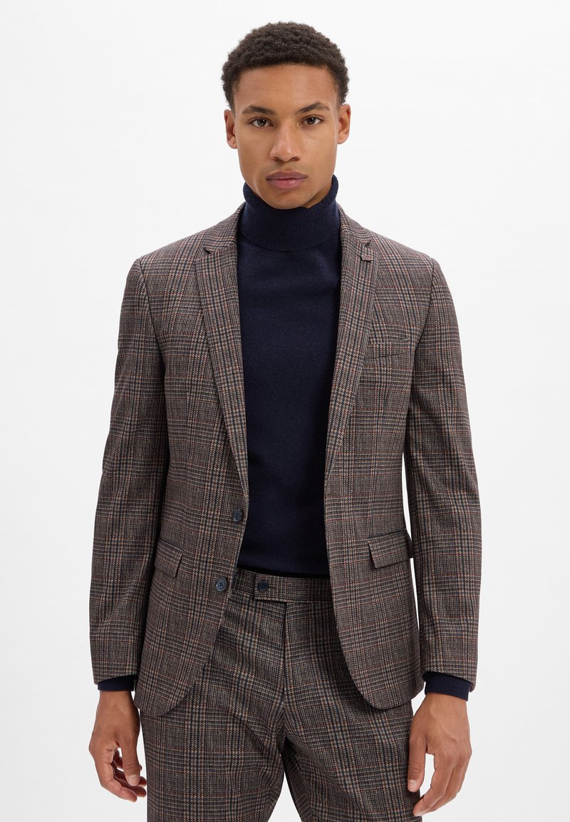 Braun kariertes Anzug mit einem strukturierten Blazer und passender Hose. Strukturierter Stoff, zwei Knöpfe und ein marineblauer Rollkragenpullover darunter.