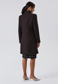 Vrouw die met haar rug naar de kijker staat, gekleed in een donkerbruin knielang jasje over een zwart geruite rok en beige hoge slingback schoenen.