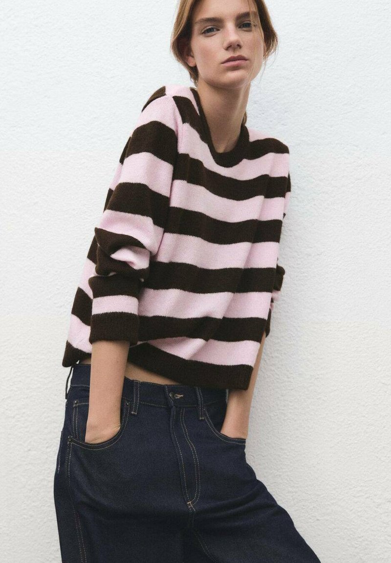 Suéter a rayas en rosa y marrón oscuro, de corte amplio con mangas largas y cuello redondeado. Combinado con jeans de mezclilla oscuros.
