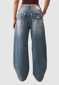 Szerokie niebieskie dżinsy z wyblakłym praniem. Posiadają dwie tylne kieszenie z dekoracyjnym przeszyciem i metalowymi akcentami.