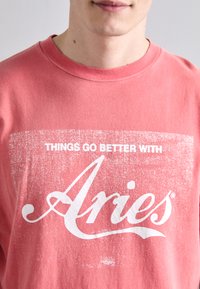 Ružová bavlnená mikina s bielym poškodeným textom, ktorý hovorí "VECI IDÚ LEPŠIE S Aries." Kulatý výstrih, mäkká textura.