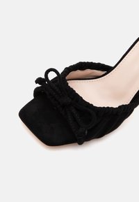 Chaussure noire à bout ouvert avec devant carré, bords froncés et un petit nœud noué sur la partie supérieure, sur fond clair.