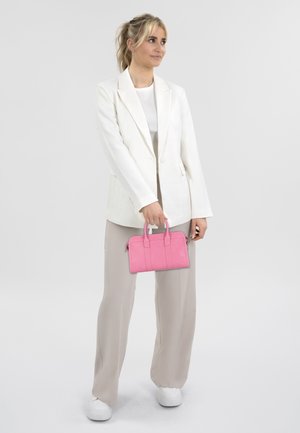 Weißer Blazer über einem hellgrauen Oberteil, kombiniert mit beigen weit geschnittenen Hosen. Hält eine leuchtend pinke Handtasche mit strukturiertem Design. Weiße Sneakers.