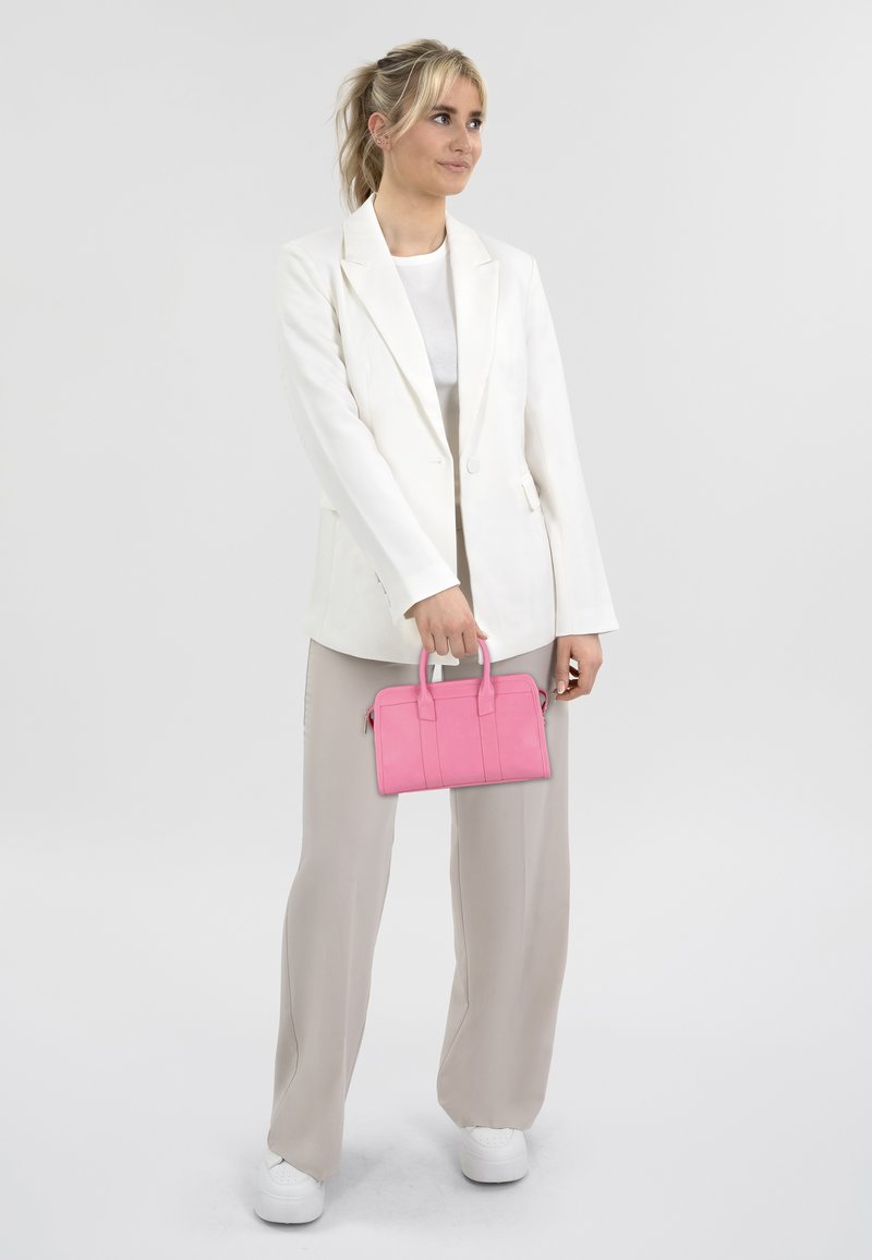 Witte blazer over een lichtgrijze top, gecombineerd met beige wijde broek. Houdt een felroze handtas met een gestructureerd ontwerp vast. Witte sneakers.