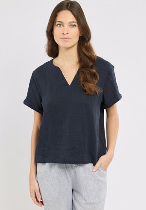 Femme aux longs cheveux bruns portant un haut texturé bleu marine à manches courtes et col en V, et un pantalon gris clair, se tenant debout avec une main dans la poche.