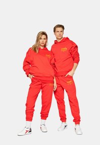 Felpe con cappuccio rosse e pantaloni jogger abbinati in un materiale morbido, con grafiche gialle, abbinate a sneakers bianche.