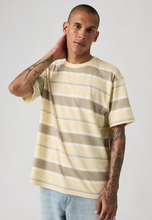 Mężczyzna z krótkimi włosami i tatuażami na rękawach, noszący żółto-beżową koszulkę w paski i jasnoniebieskie dżinsy, pozujący z ręką na szyi.