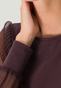 Mørklilla top med teksturerede, gennemsigtige puffærmer, glat stof og rund halsudskæring, der fremhæver en blanding af bløde og luftige materialer.