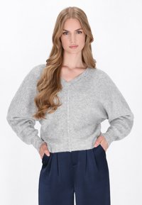Helles graues gestricktes Sweatshirt mit V-Ausschnitt und weiten Ärmeln, das ein strukturiertes Muster aufweist. Kombiniert mit dunkelblauen, gefalteten Hosen.