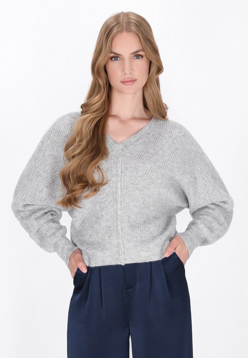 Helles graues gestricktes Sweatshirt mit V-Ausschnitt und weiten Ärmeln, das ein strukturiertes Muster aufweist. Kombiniert mit dunkelblauen, gefalteten Hosen.