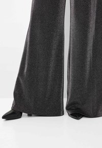 Hoge taille, glanzende zwarte wijde broek gemaakt van een textuur, metallic stof, contrasterend met puntige zwarte schoenen aan de onderzijde.