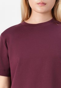 Woman wearing a plain burgundy short-sleeve crewneck t-shirt, cropped from chin to mid-torso on a white background.