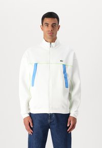 Balta zip-up striukė su mėlynomis kišenėmis, žaliais akcentais ir aukštu apykakle, pagaminta iš minkštos medžiagos. Įprasta fit'as su lygaus tekstūros.