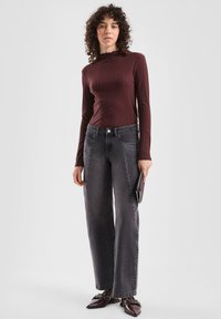 Langsleeve bordeaux ribbeltop met een gerimpelde kraag, gecombineerd met loszittende donkergrijze jeans en puntige schoenen, terwijl ze een zwarte clutch vasthoudt.