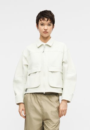 Ung person med kort, mørkt hår iført en hvid lynlåstrøje med store frontlommer og beige bukser, stående mod en ensfarvet baggrund.