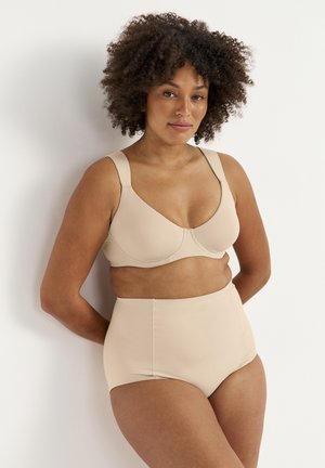 Kvinde med krøllet hår iført beige støttende BH og trusser med høj talje, stående op ad en hvid væg med hænderne bag ryggen.