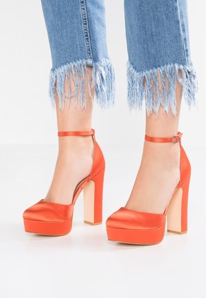 Nogi w niebieskich dżinsach z postrzępionymi nogawkami i jasnopomarańczowych satynowych platformach na wysokich obcasach z paskami wokół kostek na białej podłodze.