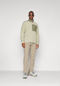 Chaqueta de forro polar en verde claro con un bolsillo en el pecho verde oscuro; combinada con pantalones beige y zapatillas gris claro. Modelo lleva gafas de sol.