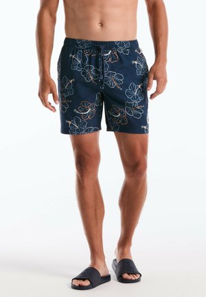 Man die marineblauwe zwembroek draagt met witte en oranje hibiscus bloemenpatronen en zwarte slidesandalen op een witte achtergrond.