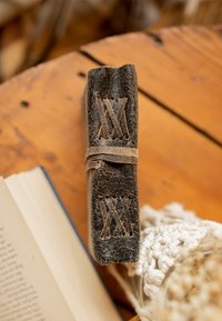 Černý kožený zápisník s viditelným šitím, s hnědou koženou sponou na zavření a s vyraženými vzory na hřbetu.