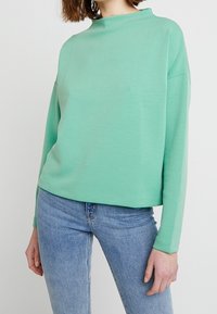 Kvinna som bär en långärmad mintgrön topp och ljusblå jeans, stående mot en vit bakgrund.