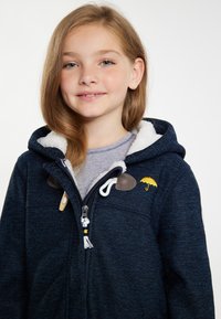 Kék kapucnis dzseki puha fehér polár béléssel, sárga esernyő hímzéssel és fa gombos díszítéssel. Cipzáras záródás zsinórral.