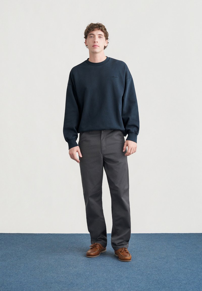 Felpa in pile blu marina, pantaloni grigi, mocassini marroni. Tessuto morbido, vestibilità comoda e design semplice. Posizione in piedi su uno sfondo con moquette blu.