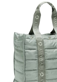 Gevoerde draagtas van lichtgroene stof, met verticale naden, korte handvaten en een contrasterende logo-ribbon voor extra details.