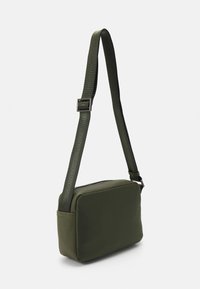Olivengrønn crossover-veske med en glatt, polstret design, justerbar reim og glidelås. Har minimal merkevare på reimen.