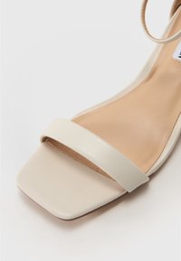 Beige nahka sandaali, jossa on neliön muotoinen varvasosa, ohut hihna jalan yli ja sileä, luonnollisen sävyinen pohjallinen. Minimalistinen muotoilu.