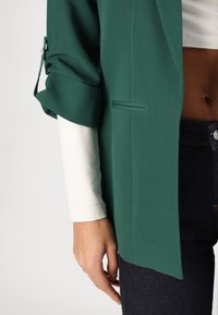 Blazer vert avec manches retroussées, présentant une texture subtile. Une petite poche à l'avant et porté sur un haut blanc à manches longues. Jean en denim foncé visible.