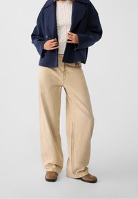 Marinblå kort jacka med stora slag, krämfärgad kabelstickad tröja, vida beige byxor och bruna ankelstövlar med platt sula.