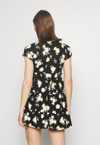 Svart blommig klänning med korta ärmar, prydd med gula blommor och gröna blad, volangkant i flera lager och justerbart midjeband.