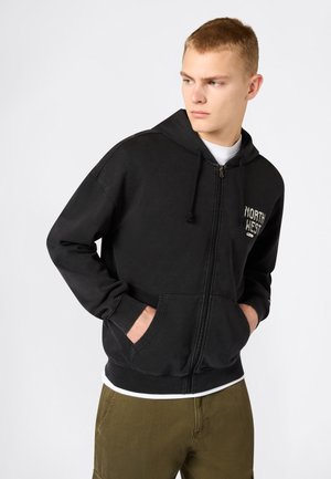 Junger Mann mit kurzen Haaren, trägt einen schwarzen Hoodie mit Reißverschluss und dem Schriftzug "NORTH WEST", Hände in den Taschen, olivegrüne Hose, schaut nach rechts.