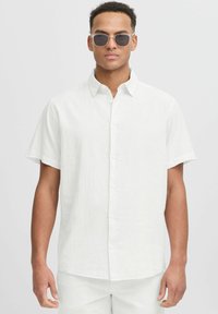 Camisa blanca de manga corta y botón, hecha de una tela texturizada, con un cuello puntiagudo y un dobladillo recto, acompañada de gafas de sol.