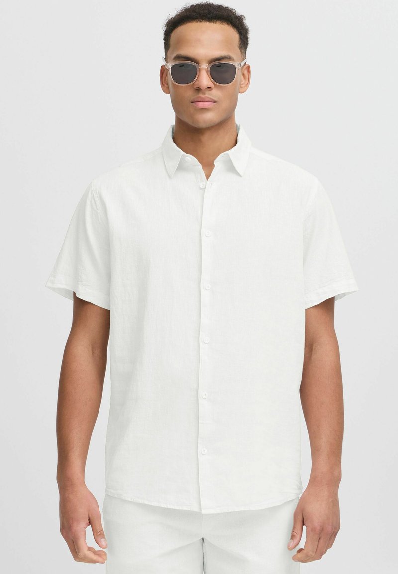 Camisa blanca de manga corta y botón, hecha de una tela texturizada, con un cuello puntiagudo y un dobladillo recto, acompañada de gafas de sol.