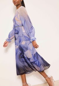 Gennemsigtig langærmet kjole med blå blomsterprint, taljeret pasform og opsmøgede ærmekanter med blomsterdetaljer, som falder til midt på læggen.