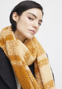Écharpe à motifs dans des teintes jaunes et beiges, avec une texture douce et un design en maille épaisse. Se drape autour du cou pour la chaleur.