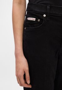 Čierne denimové džínsy s úzkym strihom, na vrecku so červenobielou nálepkou "Calvin Klein" a kovovými gombíkmi.