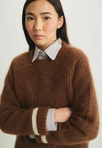 Pull en maille bouclée marron avec une finition texturée, présentant un revers rayé blanc et marron. Porté par-dessus une chemise à col rayée.