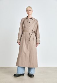 Beige esőkabát övvel a deréknál, nagyméretű gallérral és gombos záródással. Hosszú ujjak és egyenes vonal jellemzi, széles lábú farmernadrággal párosítva.
