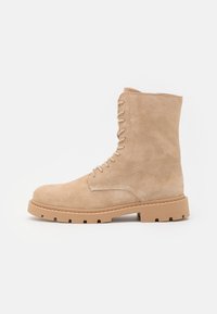 Stivale alla caviglia in suede color sabbia, con lacci, suola in gomma spessa e dettagli di cucitura, mostrato di lato su sfondo bianco.