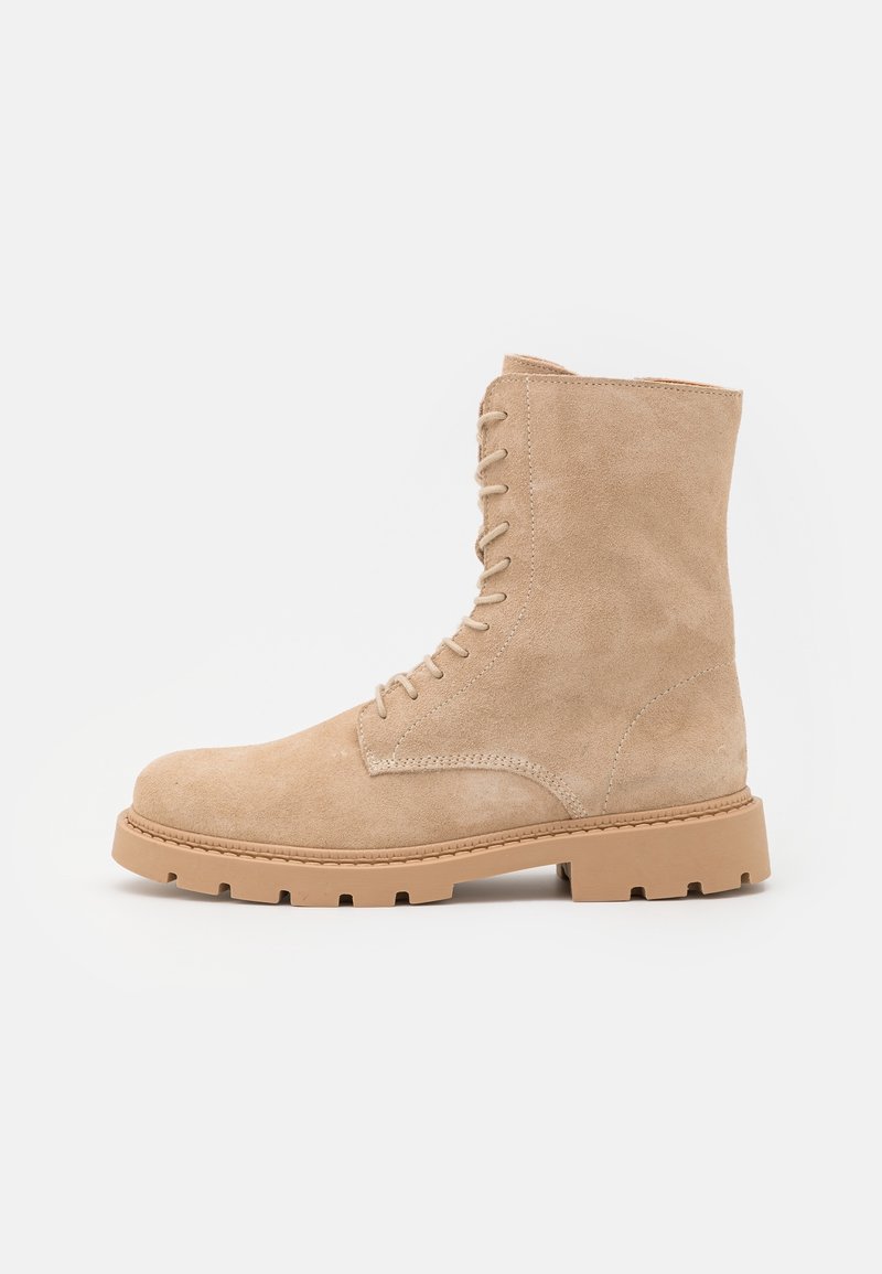 Stivale alla caviglia in suede color sabbia, con lacci, suola in gomma spessa e dettagli di cucitura, mostrato di lato su sfondo bianco.
